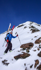 male backcountry skier on a ski mountaineering tour on his way to the summit with his skis strapped to his backpack on a gorgeous winter fay in the Alps