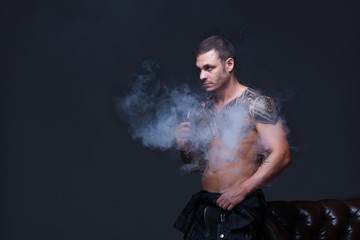 Vaper. The man with a muscular naked torso with tattoos smoke an electronic cigarette on the dark background