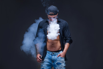 Vaper. The man dressed blue jeans, black shirt and black baseball cap with tattoos smoke an electronic cigarette on the dark background