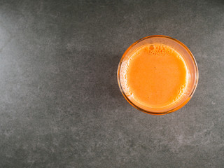Glass carrot juice and fresh carrots with green leaves around it over gray stone background.