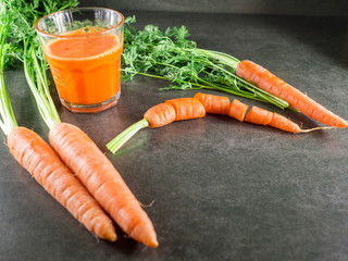 Glass carrot juice and fresh carrots with green leaves around it over gray stone background.