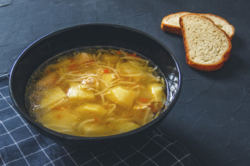 Chicken soup with potatoes, noodles, onions, carrots and two pieces of bread on a black background.