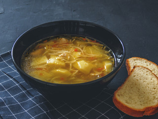 Chicken soup with potatoes, noodles, onions, carrots and two pieces of bread on a black background.