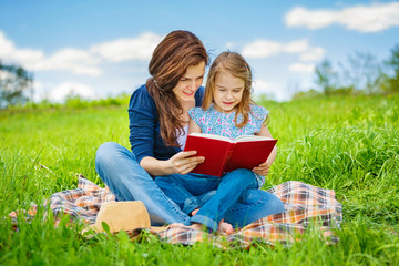 Mother and daughter enjoy reading book sitting on green meadow in the summer park