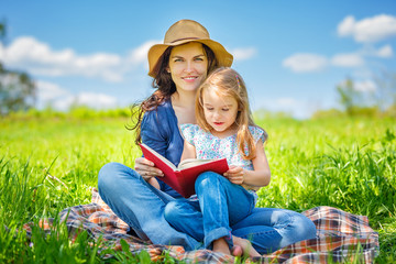 Naklejka na ściany i meble Mother and daughter enjoy reading book sitting on green meadow in the summer park