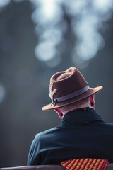 Man in brown fedora hat sitting outdoor on bench. Rear view.