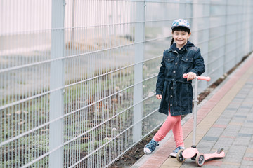 Cute little girl in jeans cardigan and on scooter on the background of the fence