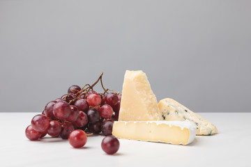 Close up view of brie, cheddar and blue cheese with grapes on gray