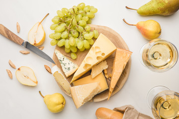 Top view of different types of cheese on wooden board surrounded by knife, fruits, almond, baguette and wine glasses on white