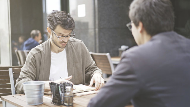 Two friends are having a conversation at table of the cafe outdoors. A man dressed in a jacket wearing eyeglasses looking into his papers in front of his friend