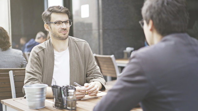 Two friends are smiling talking having a conversation at table of the cafe outdoors. A man dressed in a jacket wearing eyeglasses listening to his friend talking