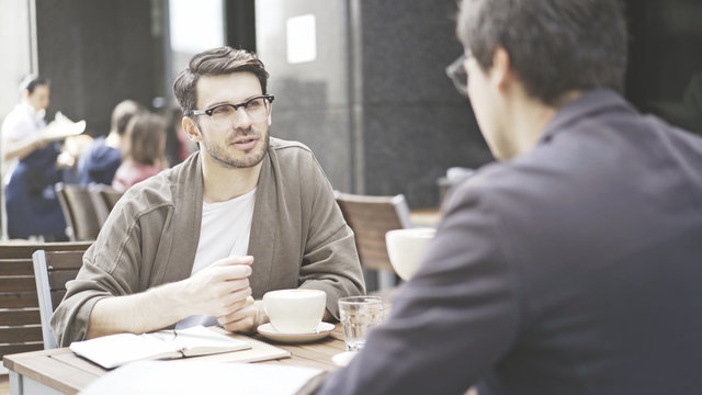 Two friends are having a conversation at table of the cafe outdoors. A man dressed in a jacket wearing eyeglasses is listening carefully to his friend talking