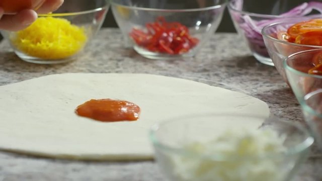 Close up shot of unrecognizable cook adding tomato sauce to future pizza and spreading it evenly with spoon, other ingredients placed around it, tracking left