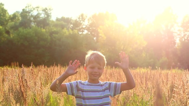 Portrait of funny little boy playing alone outside in long green and golden grass in countryside meadow on bright sunny summer evening. Child waves his hands to camera. Video shot at golden hours.