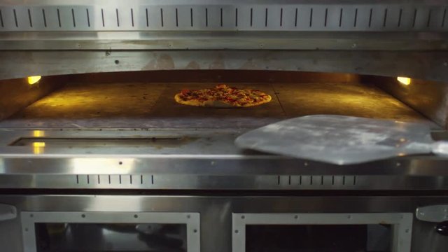 Unrecognizable cook opening oven, using peel to take pizza out of it and putting finished pizza on wooden surface