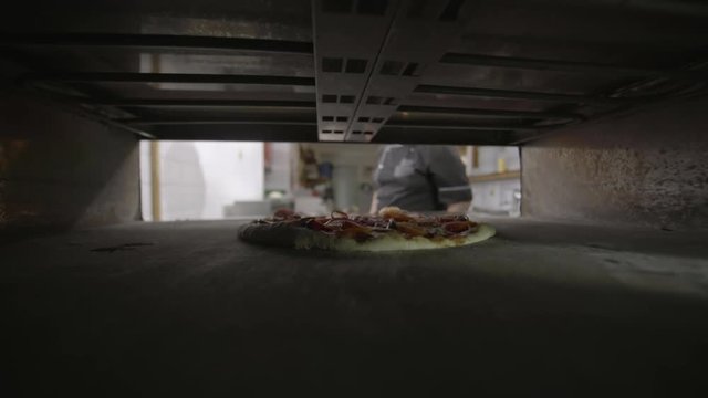Male restaurant chef baking pizza and using peel to put in oven, close up shot, view from the oven