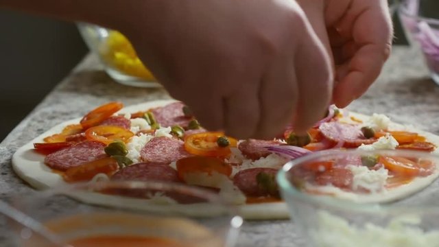 Close up shot of restaurant chef adding red onion circles to pizza topped with grated cheese, tomatoes, salami, and capers