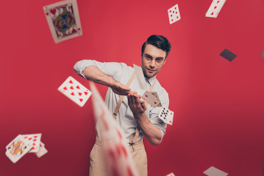 Professional, cunning joyful magician, illusionist, gambler with tricky glance in casual outfit, glasses, throwing, sending cards to the camera, standing over red background