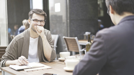 Two friends are laughing at table of the cafe outdoors. A man dressed in a jacket wearing eyeglasses is laughing smiling with his friend