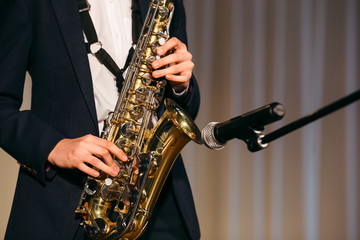 Fototapeta premium Young boy hands in black jacket and white shirt playing saxophone at the microphone on stage