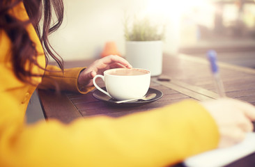 leisure and people concept - close up of young woman or teenage girl with cup drinking cocoa and writing to notebook at city street cafe terrace
