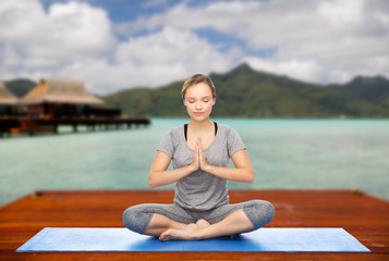 Fototapeta na wymiar fitness, meditation and healthy lifestyle concept - happy woman making yoga lotus pose on wooden pier over island beach and bungalow background
