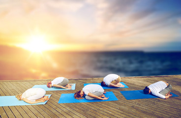 fitness and healthy lifestyle concept - group of people doing yoga child pose outdoors on wooden pier over sea background