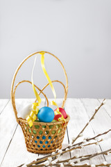 Painted bright Easter eggs in a wicker basket and willow branches on a light wooden table. Copy space
