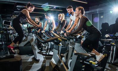 Sporty people on riding exercise bikes with assistance of their trainer during cycling class