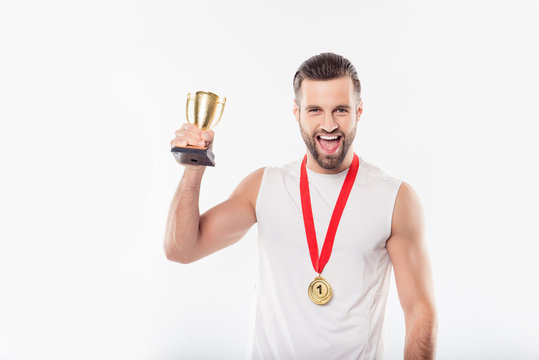 Attractive powerful sporty strong cheerful positive confident cheerful man in t-shirt having gold medal with red ribbon on his neck, showing, raising cup trophy, yelling over white background