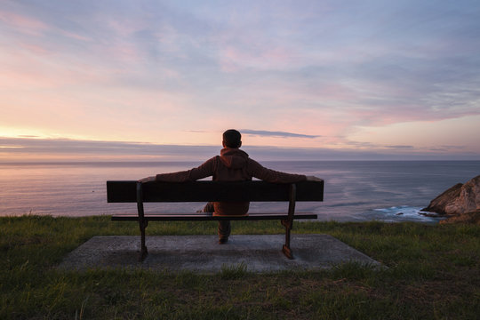Full length of man looking at sea while sitting on bench against cloudy sky during sunset
