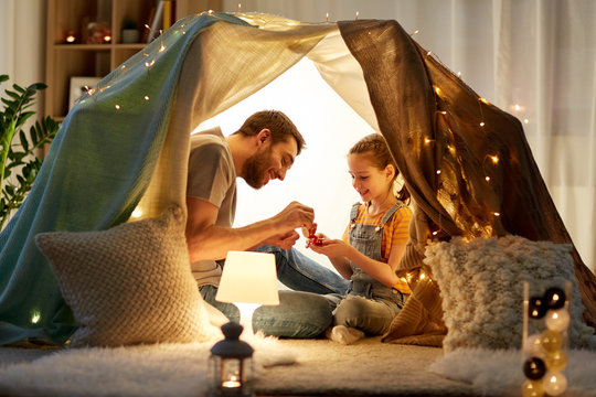 family, hygge and people concept - happy father and little daughter playing tea party in kids tent at night at home