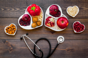 Products good for heart and blood vessels. Vegetables, fruits, nuts in heart shaped bowl near stethoscope on dark wooden background top view