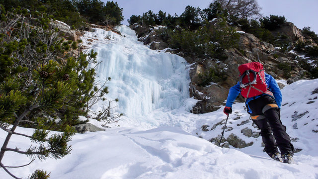male ice climber in a blue jacket and red backpack approaches an ice fall on a beautiful winter day adn prepares to climb it