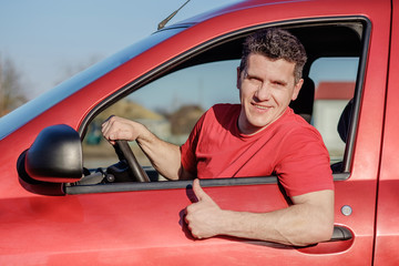 Attractive white middle aged man sitting in red car, smiling and doing gesture thumb up.