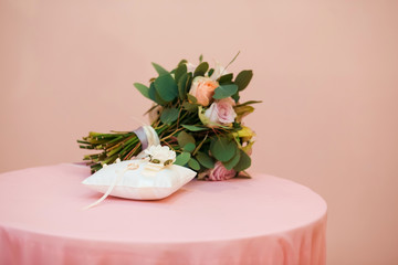 Bridal bouquet. Wedding. Beautiful bouquet with roses and calla flowers at white, pink colour, decorated with silk lilac ribbon lies on a table with pink silk fabric. Bridal rings at white pillow.