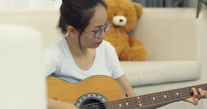 Asian young girl sitting on long sofa chair concentrating focused learning to play guitar at home.