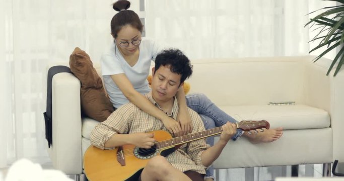 Asian man serenades sweetheart with guitar woman looks at boyfriend artistic intent shallow depth at home.