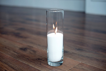 A burning candle in tall glass vase flask standing on the floor in the middle of room.