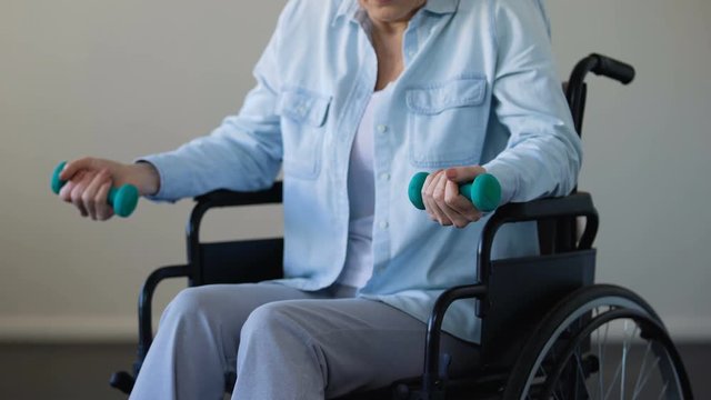 Mature lady sitting in wheelchair and trying to lift dumbbells, rehabilitation