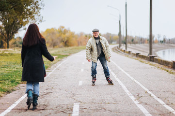 Happy old man goes on rollers with his daughter on the road in autumn park near embankment. Happy pension.