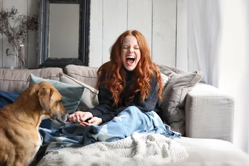 Fotobehang Hübsche rothaarige Frau sitzt lachend auf einer Couch während ein Hund an ihrer Hand schnüffelt © Joerch