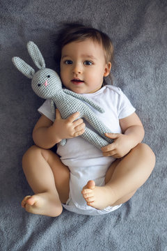 very cheerful baby boy in white clothes lying on the bed with a knitted toy rabbit