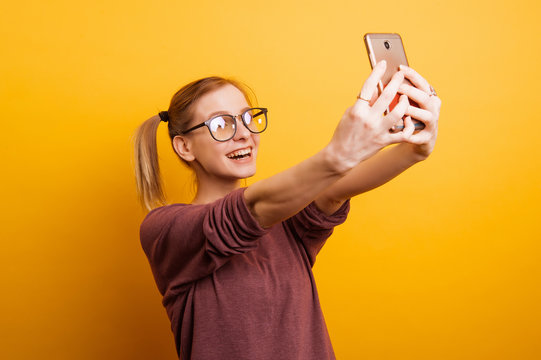 Selfie time. Joyful young blonde women with eye glasses making selfie by her smart phone isolated over yellow background.