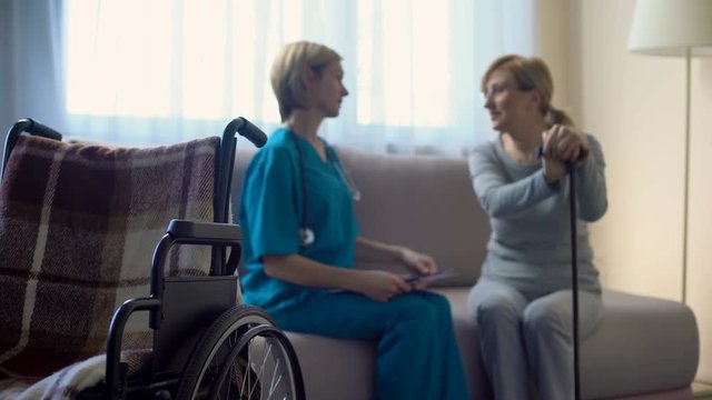 Nurse and aged woman sitting on sofa at hospital, discussing news, support