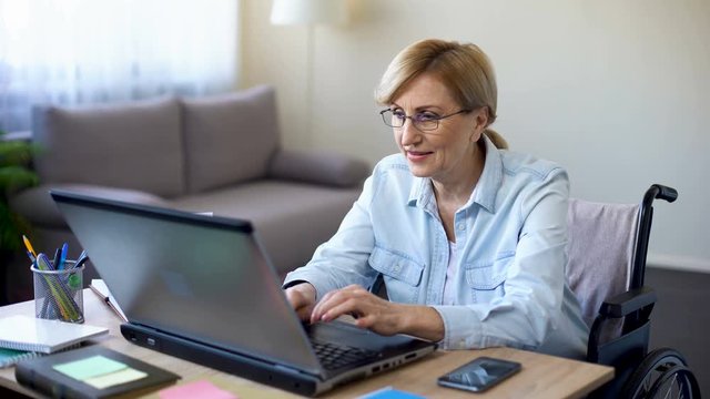 Successful senior woman in wheelchair working on laptop, chatting with client