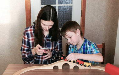 Playing together. Mom looks at phone and son is playing a wooden railway with train, wagons and tunnel sitting at the table.