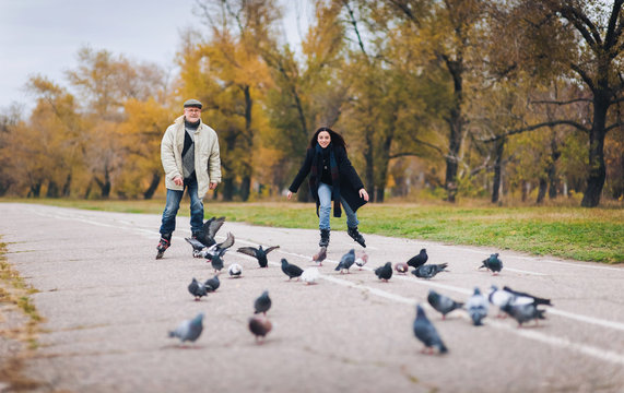An original elderly man and his pretty daughter have fun skating on roller skates in autumn park. People and pigeons.