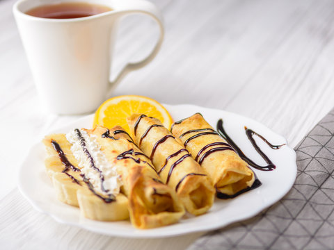Pancakes with banana, whipped cream decorated with chocolate syrup on white wooden background. And a cup of tea