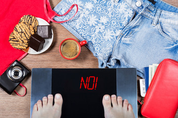 Woman weighs herself before vacation. Concept of unhealthy lifestyle and its consequenses. Digital scales with sign no! and holiday accessories and sweets. Top view